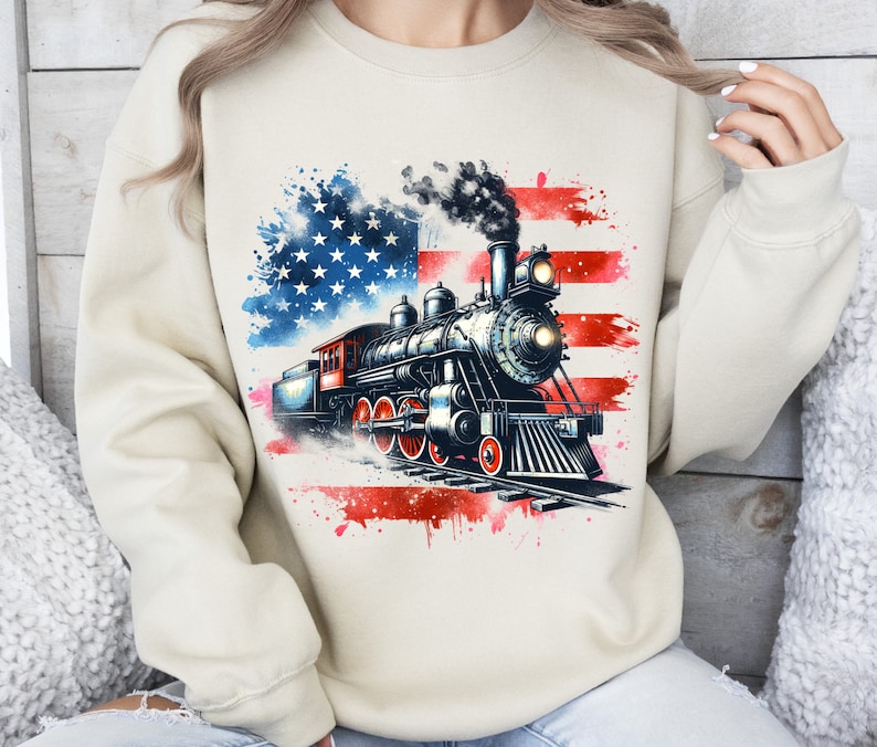 May include: A beige sweatshirt with a watercolor American flag design featuring a vintage steam locomotive. The locomotive is black and red with white smoke coming from the chimney.