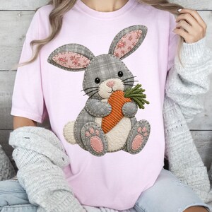Pode incluir: Uma t-shirt rosa claro com um gráfico central de um coelho cinzento aos quadrados com barriga creme e almofadas cor-de-rosa nas patas. O coelho segura uma cenoura laranja texturizada com folhas verdes. As orelhas têm um padrão floral rosa.