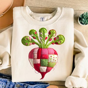 Puede incluir: Sudadera de color crema con un diseño de remolacha colorido al estilo patchwork. La remolacha tiene una combinación de colores rojo, rosa y verde con elementos detallados de hojas y tallos. El diseño está centrado en la parte delantera.