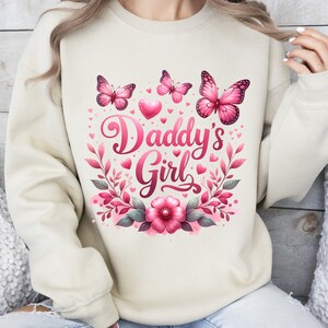 May include: A cream-colored sweatshirt with a pink floral design and the text "Daddy's Girl". The design includes butterflies, hearts, and leaves.