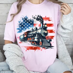 May include: A pink t-shirt with a watercolor design of an American flag and a vintage steam locomotive. The locomotive is black and gray with red accents.