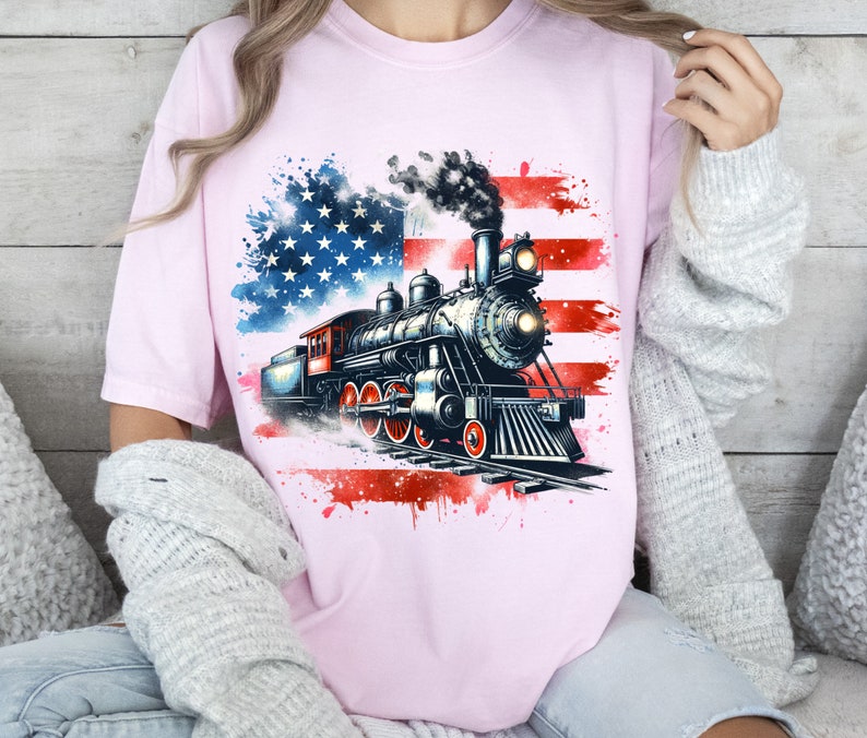 May include: A pink t-shirt with a watercolor design of an American flag and a vintage steam locomotive. The locomotive is black and gray with red accents.