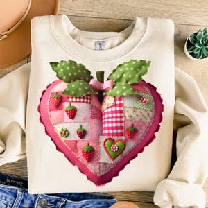 May include: Cream-colored sweatshirt featuring a large, heart-shaped strawberry design. The strawberry is made of patchwork fabric in pink, red, and green, with embroidered strawberries and leaves. A cute and cozy design for a casual look.