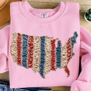 May include: A light pink sweatshirt featuring a decorative map of the United States. The map design incorporates red, white, and blue fabric strips, creating a patriotic aesthetic. The sweatshirt is made of a soft material.