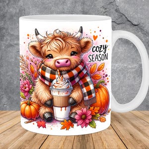 Puede incluir: Taza de cerámica blanca con una vaca de las Tierras Altas de dibujos animados con una bufanda a cuadros, sosteniendo una bebida de café. La taza tiene las palabras "cozy SEASON" y está decorada con calabazas, flores y hojas de otoño.