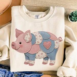 May include: A cream-colored crewneck sweatshirt featuring a large graphic of a pig composed of various fabric-style patches. The pig design includes pink and blue solid colors mixed with floral patterns, a small pink heart on its side, and a curly tail.