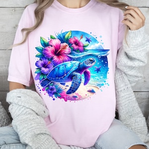 May include: A pink t-shirt with a watercolor design of a sea turtle swimming in the ocean with tropical flowers. The turtle is blue and green with a brown shell. The flowers are pink, purple, and blue.