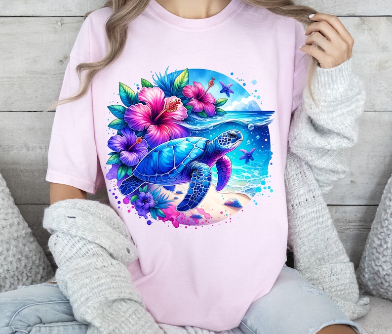 May include: A pink t-shirt with a watercolor design of a sea turtle swimming in the ocean with tropical flowers. The turtle is blue and green with a brown shell. The flowers are pink, purple, and blue.