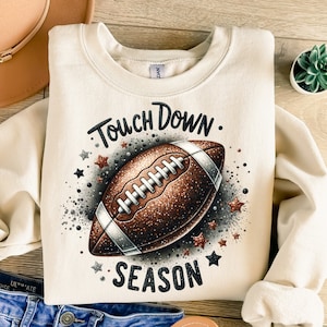 May include: A cream-colored sweatshirt with a brown football graphic and the text "Touch Down. Season" in black lettering.