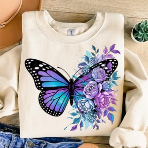 May include: Cream-colored sweatshirt featuring a large butterfly design with purple, blue, and black wings. The butterfly is surrounded by purple and blue flowers and foliage. The sweatshirt has a crew neck and long sleeves.
