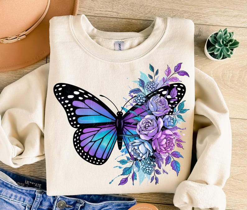May include: Cream-colored sweatshirt featuring a large butterfly design with purple, blue, and black wings. The butterfly is surrounded by purple and blue flowers and foliage. The sweatshirt has a crew neck and long sleeves.