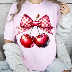 May include: Pink t-shirt with a graphic of two red cherries tied together with a pink and white polka dot bow.