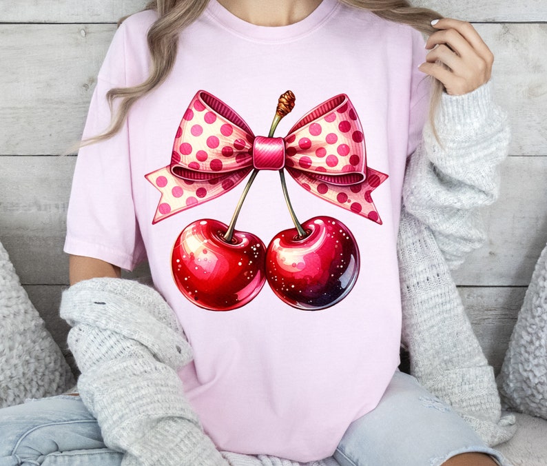 May include: Pink t-shirt with a graphic of two red cherries tied together with a pink and white polka dot bow.