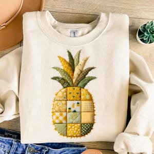 May include: Cream-colored sweatshirt featuring a detailed pineapple design. The pineapple is composed of various fabric patterns in shades of yellow, green, and tan. The design is centered on the front of the long-sleeved top.