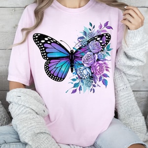 May include: A light pink t-shirt featuring a large butterfly design with purple and teal wings, and black accents. The butterfly is surrounded by purple and blue flowers and foliage. The shirt is a casual, everyday wear item.