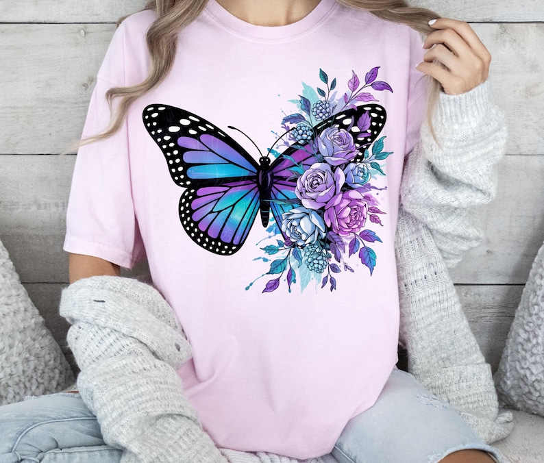 May include: A light pink t-shirt featuring a large butterfly design with purple and teal wings, and black accents. The butterfly is surrounded by purple and blue flowers and foliage. The shirt is a casual, everyday wear item.