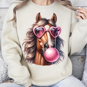 May include: A beige sweatshirt featuring a graphic of a brown horse wearing pink heart-shaped sunglasses and blowing a pink bubble.