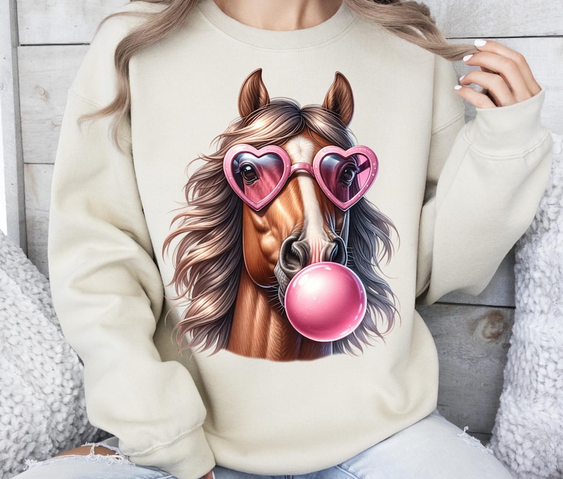 May include: A beige sweatshirt featuring a graphic of a brown horse wearing pink heart-shaped sunglasses and blowing a pink bubble.