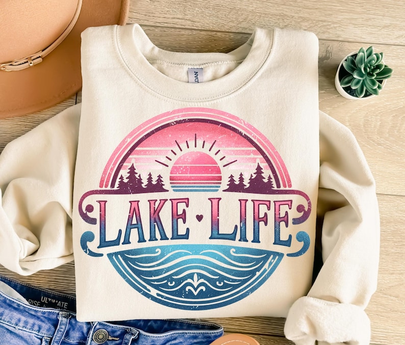 May include: A cream-coloured sweatshirt with a vintage-style graphic of a sunset over a lake. The graphic features a pink and blue colour scheme and the text "LAKE LIFE" in a bold, stylised font.