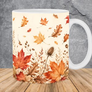 May include: A white ceramic mug featuring a wrap-around print of autumn foliage, including various maple and oak leaves in shades of orange, red, and brown, interspersed with dried thistle and delicate wild grasses on a cream-colored background.