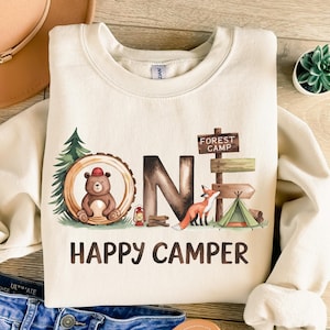 May include: A cream-colored sweatshirt with a graphic of a bear, fox, and tent in a forest setting. The graphic says "One Happy Camper" and includes a sign that says "Forest Camp".