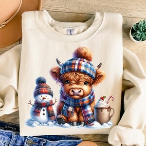 May include: Cream-colored sweatshirt featuring a Highland cow wearing a plaid hat and scarf, with a snowman and a mug of hot chocolate. The cow has brown fur and is surrounded by snow. A cozy winter-themed design.