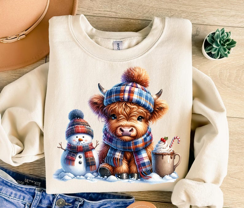 May include: Cream-colored sweatshirt featuring a Highland cow wearing a plaid hat and scarf, with a snowman and a mug of hot chocolate. The cow has brown fur and is surrounded by snow. A cozy winter-themed design.