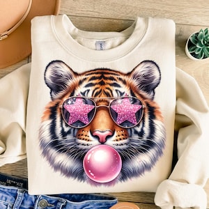 May include: A cream-coloured sweatshirt featuring a graphic of a tiger wearing pink star-shaped sunglasses and blowing a pink bubble.