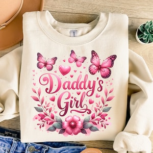 May include: A cream-colored sweatshirt with a pink floral design and the text "Daddy's Girl" in pink lettering. The design includes butterflies, hearts, and leaves.