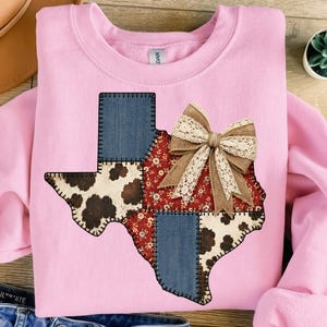 May include: A light pink sweatshirt featuring a patchwork design of the state of Texas. The Texas outline is composed of denim, floral, and cowhide patterns, with a burlap and lace bow. The sweatshirt is a casual style.
