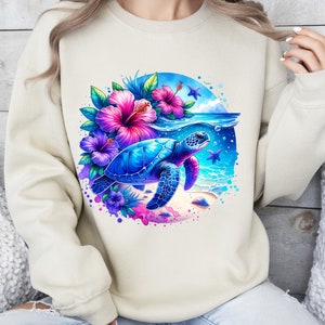 May include: A cream colored sweatshirt with a colorful graphic of a sea turtle swimming in the ocean with pink and purple flowers. The graphic is surrounded by a blue circle with white splashes of paint.