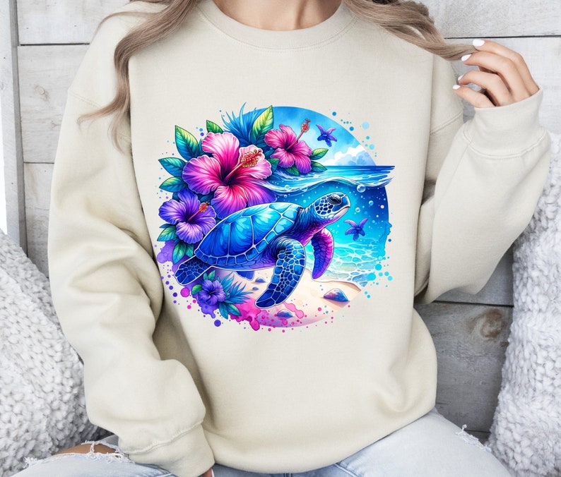 May include: A cream colored sweatshirt with a colorful graphic of a sea turtle swimming in the ocean with pink and purple flowers. The graphic is surrounded by a blue circle with white splashes of paint.