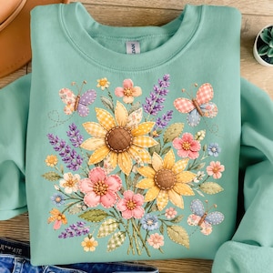 Puede incluir: Una sudadera de color verde azulado claro con un diseño floral con girasoles, flores rosas y azules y mariposas. Las flores y las mariposas tienen una apariencia acolchada. La sudadera tiene cuello redondo y mangas largas.