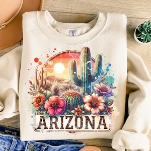 May include: A beige sweatshirt featuring a colourful graphic print of a cactus, flowers, and a sunset. The graphic also includes the text "ARIZONA" in a vintage-style font.