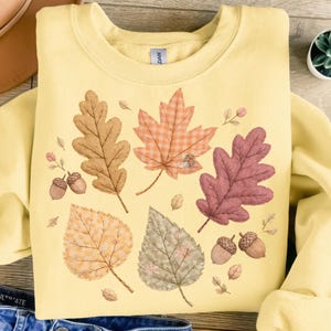 May include: A pale yellow sweatshirt with a seasonal autumn design. The design features leaves in brown, orange, and purple, with acorns and small floral details. The leaves have a quilted appearance.