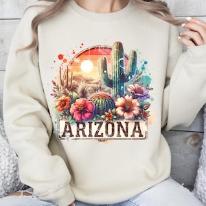 May include: A beige sweatshirt featuring a watercolour design of a cactus, flowers, and a sunset. The design includes the word "Arizona" in a vintage-style font.