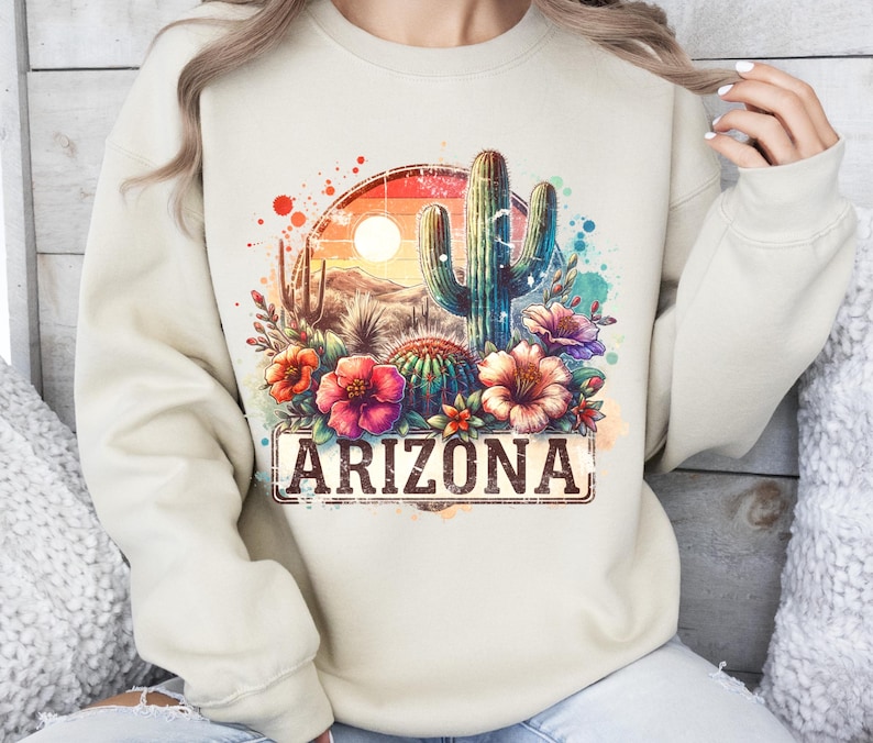 May include: A beige sweatshirt featuring a watercolour design of a cactus, flowers, and a sunset. The design includes the word "Arizona" in a vintage-style font.