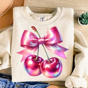 Könnte beinhalten: Ein cremefarbener Pullover mit einer pinkfarbenen, schimmernden Schleife und zwei Kirschen mit schimmernden Highlights.