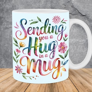 May include: White ceramic mug with the text "Sending you a Hug in a Mug" in colorful watercolor lettering. The mug is decorated with floral accents in pink, yellow, and green. The mug has a white handle and is set against a wooden surface.