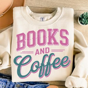 Pu&ograve; includere: Felpa color crema con le parole "BOOKS AND COFFEE" in lettere rosa e turchese. La felpa &egrave; piegata su una superficie di legno. Un cappello e jeans sono parzialmente visibili sullo sfondo.