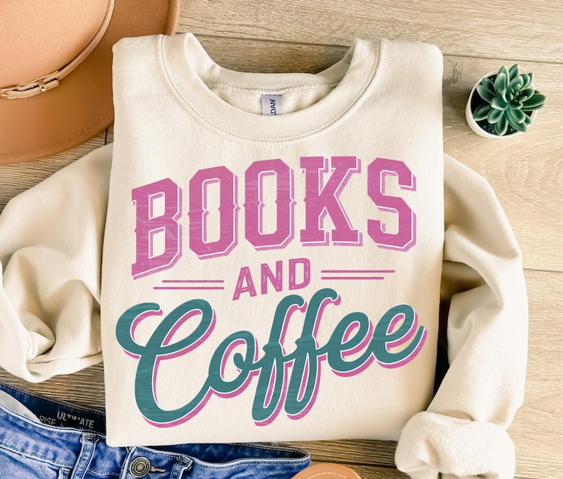 Pu&ograve; includere: Felpa color crema con le parole "BOOKS AND COFFEE" in lettere rosa e turchese. La felpa &egrave; piegata su una superficie di legno. Un cappello e jeans sono parzialmente visibili sullo sfondo.