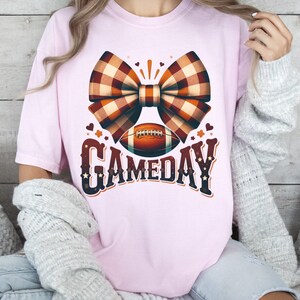 May include: A pink t-shirt with a brown and white plaid bow and a football with the text "GAMEDAY" in brown and white.