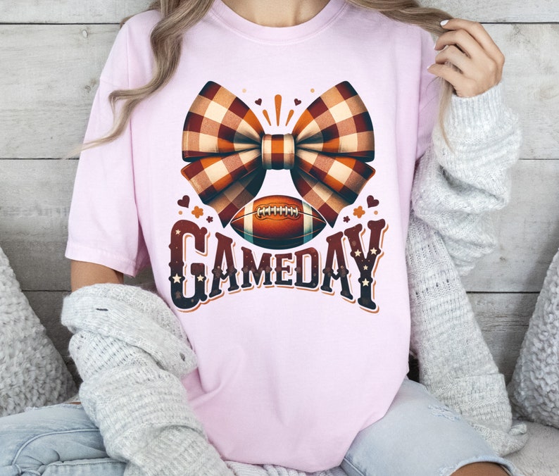May include: A pink t-shirt with a brown and white plaid bow and a football with the text "GAMEDAY" in brown and white.