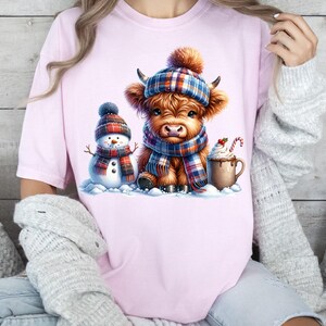 May include: Pink t-shirt featuring a watercolor illustration of a Highland cow wearing a plaid hat and scarf, a snowman, and a mug of hot chocolate. The cow and snowman are in a snowy scene, creating a cozy winter theme.