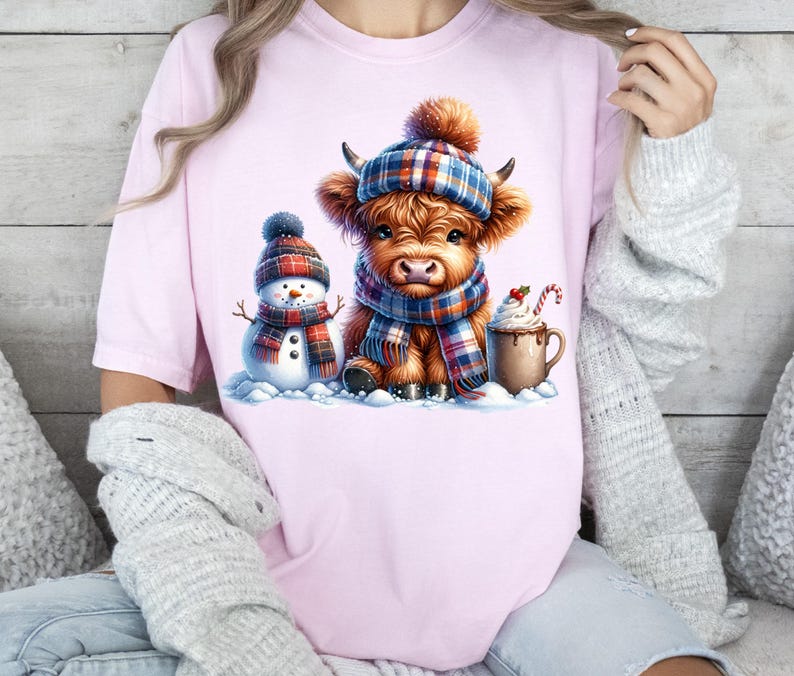 May include: Pink t-shirt featuring a watercolor illustration of a Highland cow wearing a plaid hat and scarf, a snowman, and a mug of hot chocolate. The cow and snowman are in a snowy scene, creating a cozy winter theme.