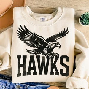 May include: A cream-colored sweatshirt with a black eagle graphic and the word "HAWKS" in black lettering.