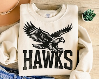 Könnte beinhalten: Ein cremefarbener Pullover mit einem schwarzen Adler-Motiv und dem Wort "HAWKS" in schwarzen Buchstaben.