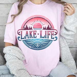 May include: A pink t-shirt featuring a vintage-style graphic of a lake scene with a sunset, trees, and the text "LAKE LIFE".