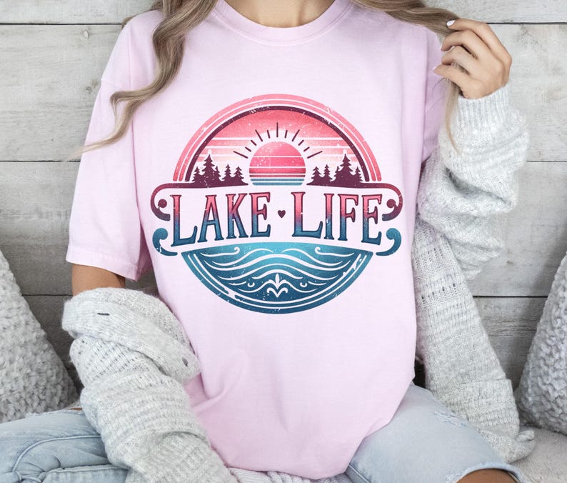 May include: A pink t-shirt featuring a vintage-style graphic of a lake scene with a sunset, trees, and the text "LAKE LIFE".