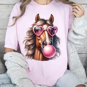 May include: A pink t-shirt featuring a graphic of a brown horse wearing pink heart-shaped sunglasses and blowing a pink bubble.